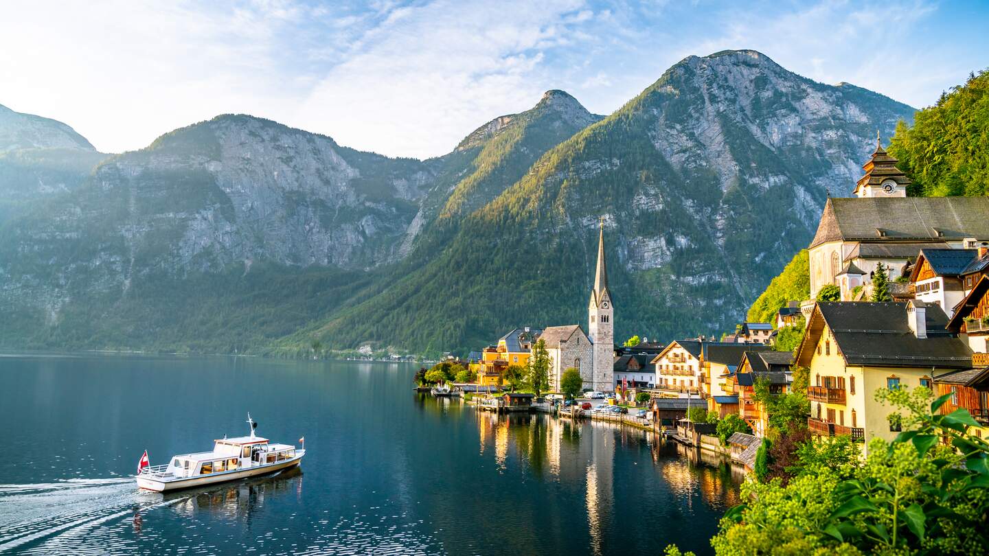 Panoramablick auf die berühmte Alte Hallstatt und den alpenländischen tiefblauen See mit Touristenschiff im malerischen goldenen Morgenlicht | © Gettyimages.com/eugen-b