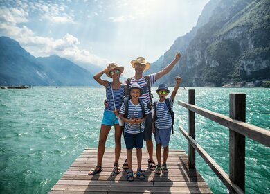 Familie genießt Urlaub in Italien. Vater und Kinder stehen auf einem Pier in Riva del Garda und jubeln in die Kamera. Dahinter bietet sich ein herrlicher Blick auf den Gardasee, umgeben von den Alpen | © Gettyimages.com/Imgorthand