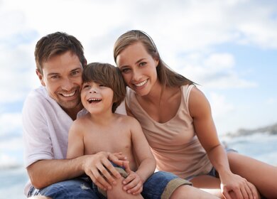 Familie am Strand | © Gettyimages.com/PeopleImages Die Eltern sitzen mit ihrem Sohn am Strand und umarmen sich | © Gettyimages.com/PeopleImages