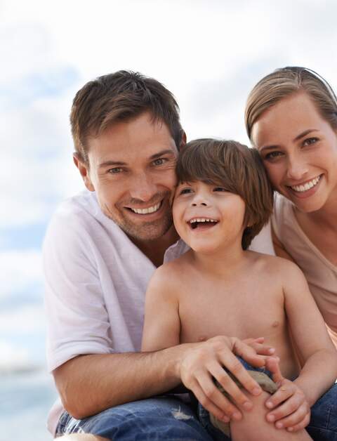 Familie am Strand | © Gettyimages.com/PeopleImages Die Eltern sitzen mit ihrem Sohn am Strand und umarmen sich | © Gettyimages.com/PeopleImages