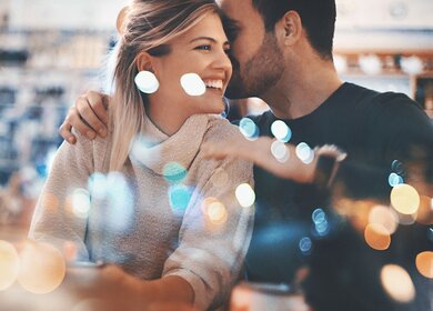 Paar auf ein romantisches Date in einem Kaffeehaus | © Gettyimages.com/gilaxia Nahaufnahme eines Paares Mitte 20, das einen romantischen Abend in einem gemütlichen Café verbringt und Spaß hat. Er flüstert ihr etwas Süßes ins Ohr und das bringt sie zum Lächeln. Von außen durch das große Fenster fotografiert, neben dem sie sitzen. Viele defokussierte, schimmernde Lichter im Rahmen. | © Gettyimages.com/gilaxia
