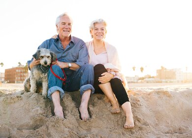 Seniorenpaar mit Hund | © Gettyimages.com/Weekend Images Inc. Seniorenpaar sitzt mit Hund im Sand am Strand | © Gettyimages.com/Weekend Images Inc.