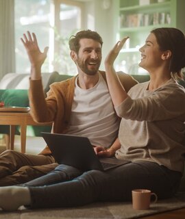 Paar gibt sich High Five | ©  Gettyimages.com/gorodenkoff Paar benutzt Laptopund gibt sich ein High-Five, während sie im Wohnzimmer auf dem Fussboden sitzen | ©  Gettyimages.com/gorodenkoff