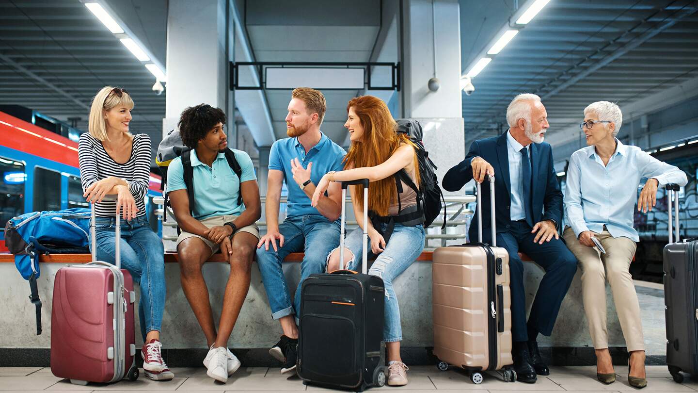 Nahaufnahme der Frontansicht einer Gruppe junger Erwachsener und eines aelteren Paares, die auf eine Zugfahrt an einem unterirdischen Bahnhof warten. Sie sitzen auf einer Bank und unterhalten sich. | © Gettyimages.com/gilaxia