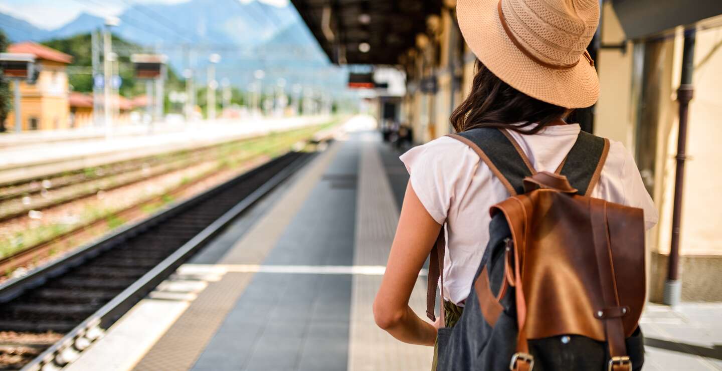 Eine Frau steht mit Strohhut und Rucksack an einem Bahnsteig im Sommer bei Sonnenschein | © MStudioImages/gettyimages.com