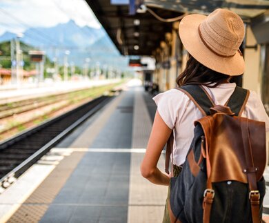 Eine Frau steht mit Strohhut und Rucksack an einem Bahnsteig im Sommer bei Sonnenschein | © MStudioImages/gettyimages.com