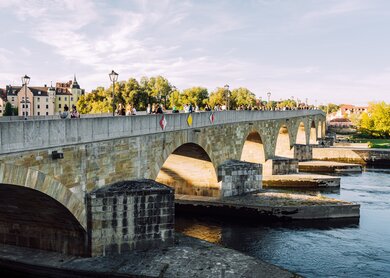 Blick auf die historische Steinbrücke in Regensburg, Bayern, Deutschland. Touristen laufen unter strahlend blauem Himmel über die Brücke. | © Gettyimages.com/Nikada
