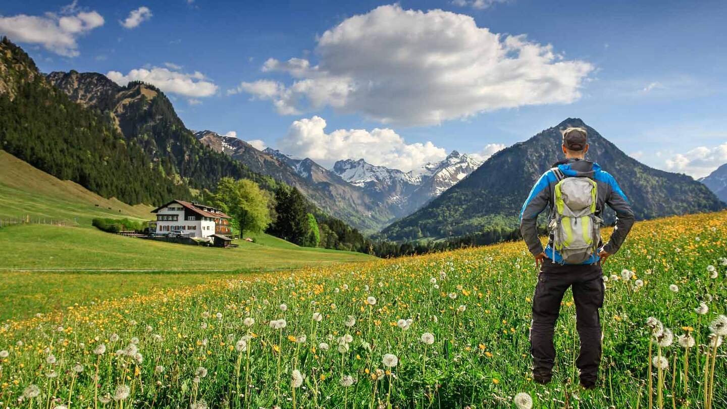Ein Wanderer mit Rucksack steht auf einer Blumenwiese, umgeben von schneebedeckten Bergen und einem traditionellen Haus in Bayern, den Alpen, Oberstdorf im Allgäu, Deutschland | © Gettyimages.com/Drepicter