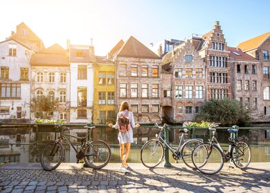Eine Frau steht in Gent, Belgien an einem Fluss und blickt auf die Altstadt. Neben ihr stehen Fahrräder | © © Gettyimages.com/RossHelen