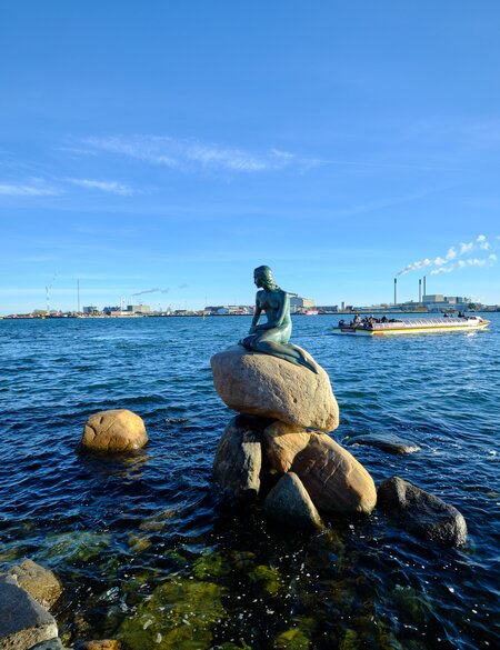 Statue der kleinen Meerjungfrau in Kopenhagen mit Touristenboot und Verbrennungsanlage im Hintergrund | © Gettyimages.com/Angelafoto