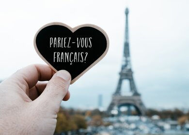 Nahaufnahme einer Hand, die ein herzförmiges Schild mit der Frage hält, ob Sie Französisch sprechen, das darin auf Französisch geschrieben ist, vor dem Eiffelturm in Paris, Frankreich | © Gettyimages.com/nito100