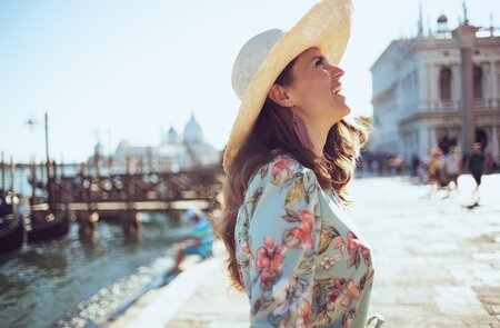 Glückliche Touristin im Blumenkleid mit Hut, die an einem sonnigen Tag in Venedig am Ufer flaniert.  | © Gettyimages.com/CentralITAlliance