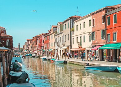 Bunte Häuser auf der Insel Murano, die an einem kleinen Kanal stehen. Touristen flanieren entlang des Kanals an einem sonnigen Tag. | © Gettyimages.com/Gavin Guan