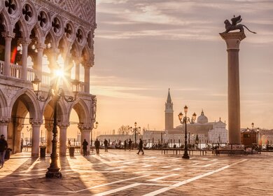 Auf dem Markusplatz in Venedig laufen Menschen. Die Sonne scheint durch den Dogenpalast. | © Gettyimages.com/tunart