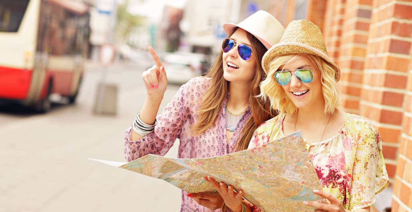 Zwei Freundinnen mit Sonnenbrillen und Hüten, die eine Karte in der Stadt benutzen | © Gettyimages.com/macniak