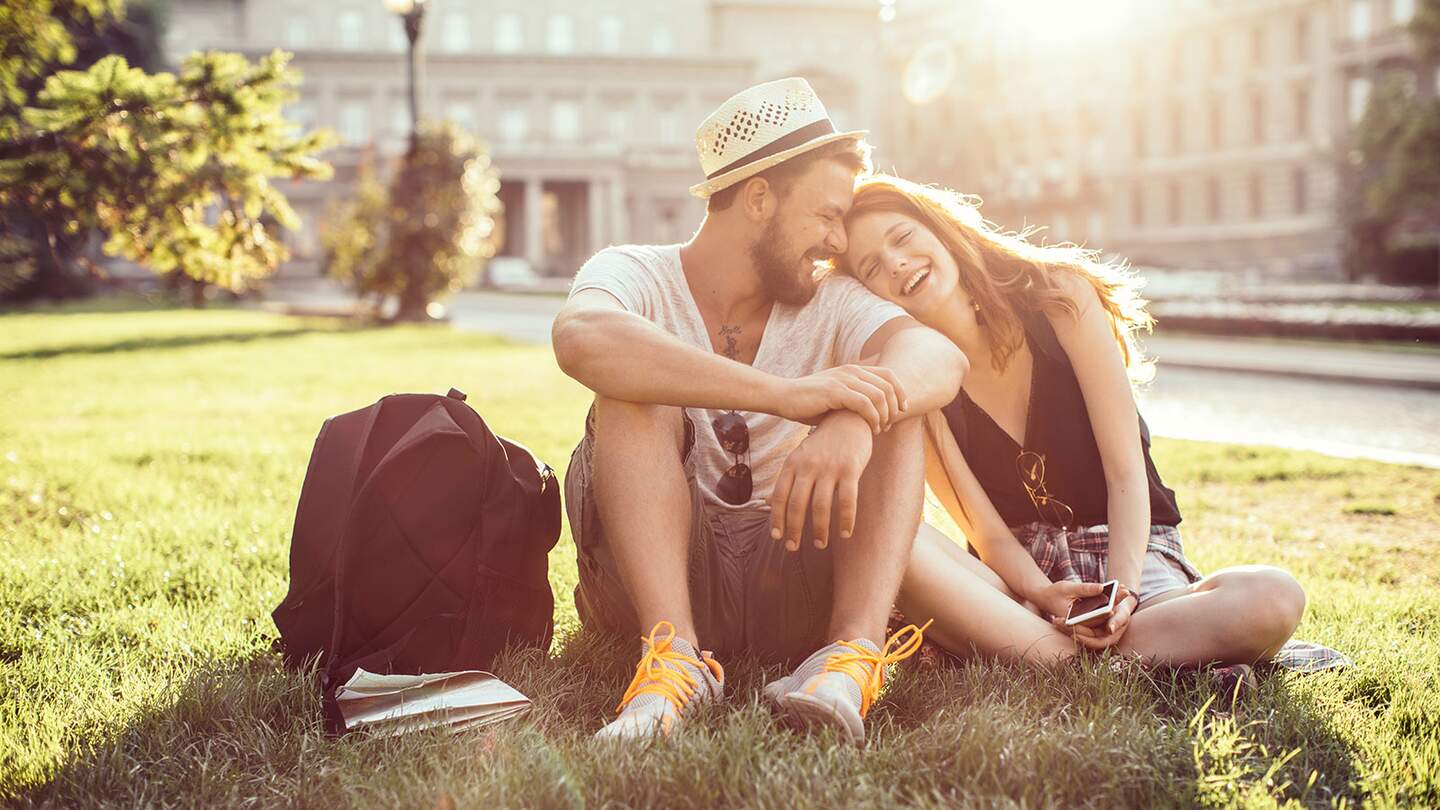 Junges Touristenpaar, das eine fremde Stadt erkundet. Im Park auf Gras sitzen und die Karte auf dem Smartphone überprüfen. Tragen Sie Freizeitkleidung und Rucksäcke. Genießen Sie den schönen sonnigen Tag. | © Gettyimages.com/svetikd