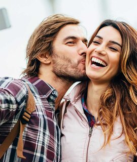 Glueckliches Paar, das ein Selfie macht, waehrend der Mann seiner Partnerin einen Kuss auf die Wange gibt | © Gettyimages.com/pixelfit