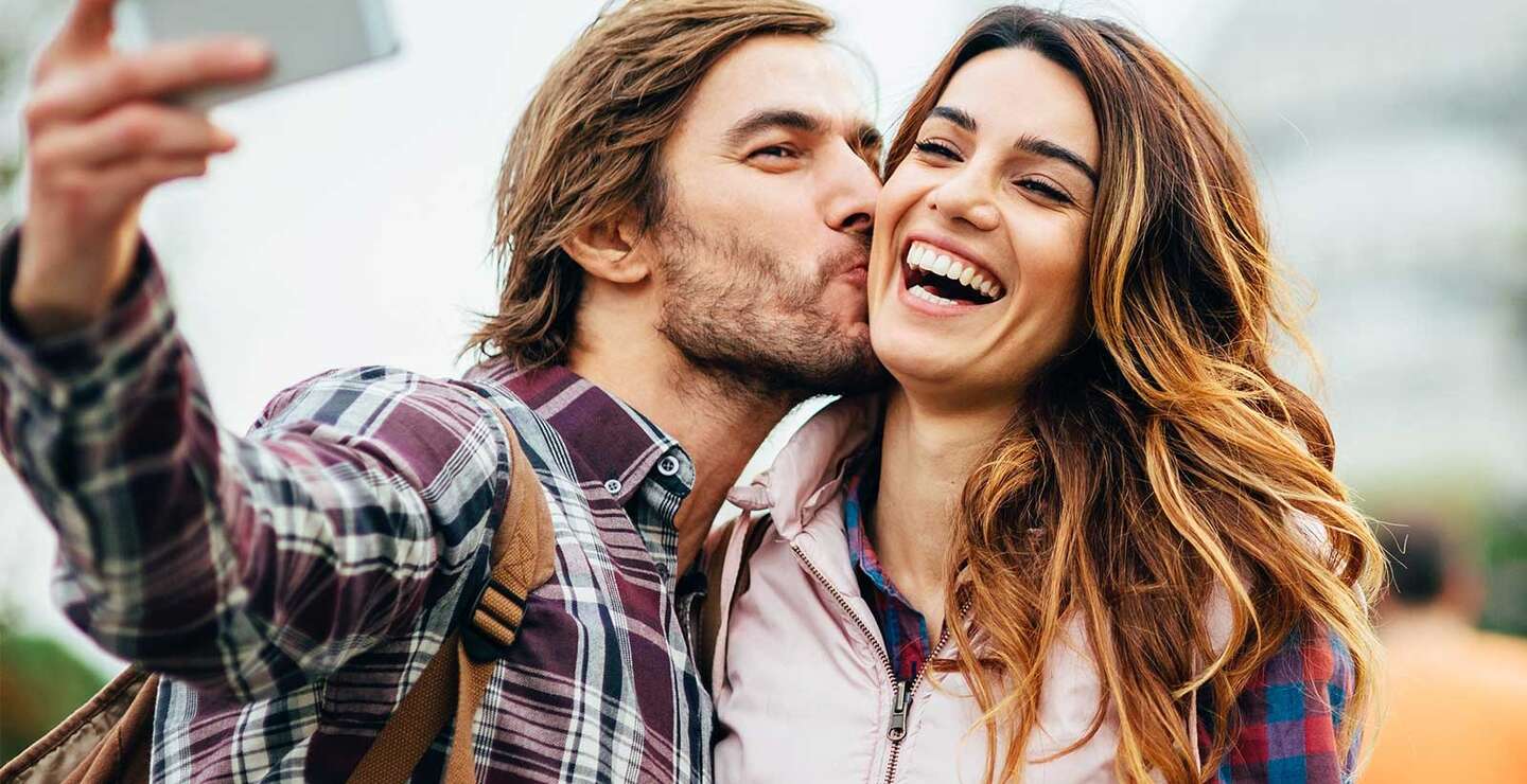 Glueckliches Paar, das ein Selfie macht, waehrend der Mann seiner Partnerin einen Kuss auf die Wange gibt | © Gettyimages.com/pixelfit