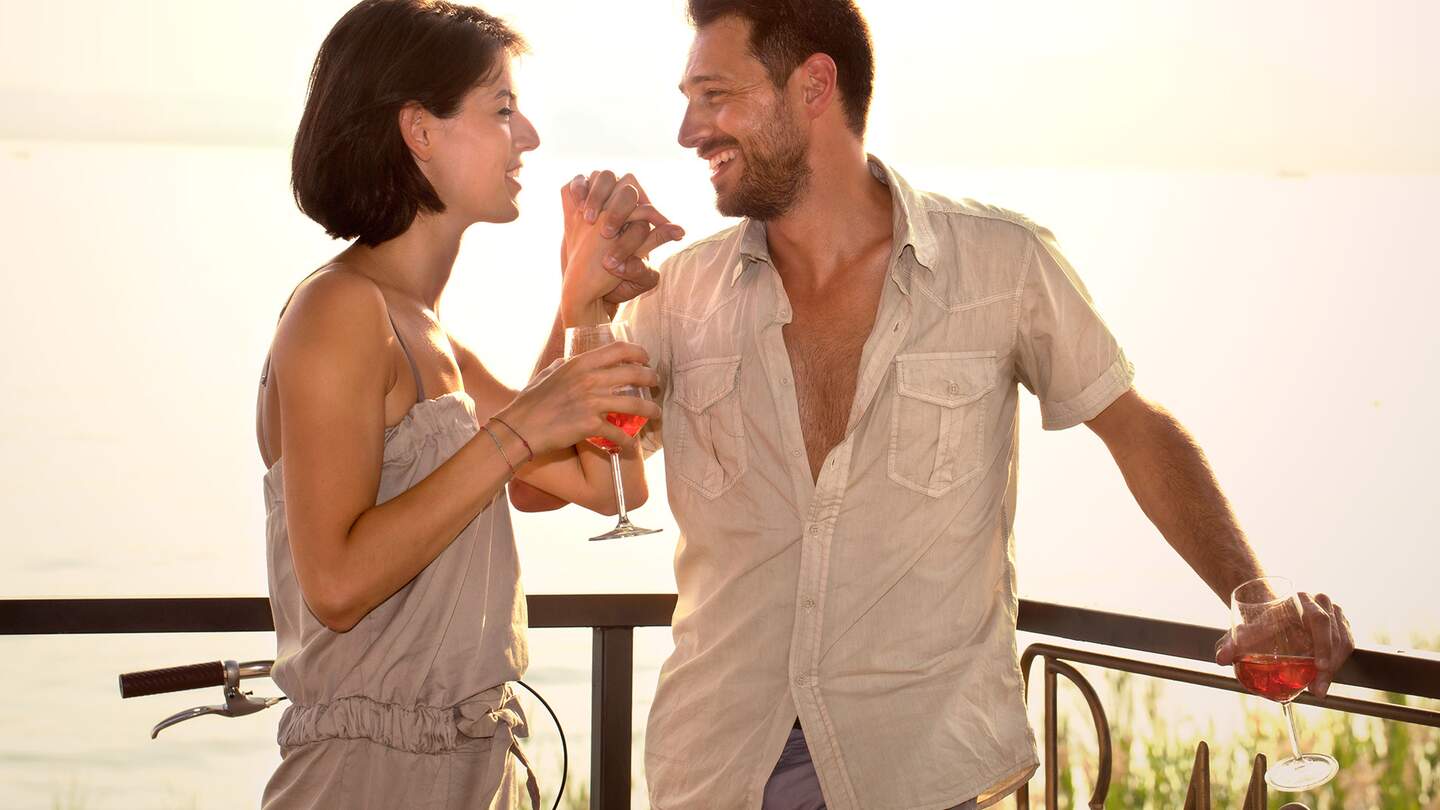 Paar steht auf einer Terrasse mit Blick auf einen See und genießt einen Aperol Spritz | © Gettyimages.com/fcsafeine