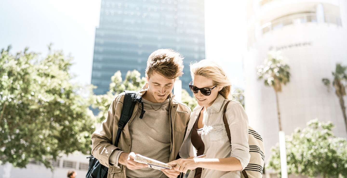Ein Foto von einem Paar, das an sonnigen Tagen in der Stadt eine Karte liest. Glückliche Touristen, die vor Gebäude stehen | ©  Gettyimages.com/tommL