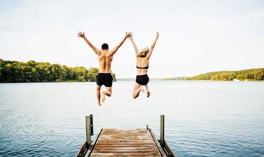 Paar das mit ihren Armen in der Luft vom Ende eines Stegs an einem See springt | © Gettyimages.com/TommL