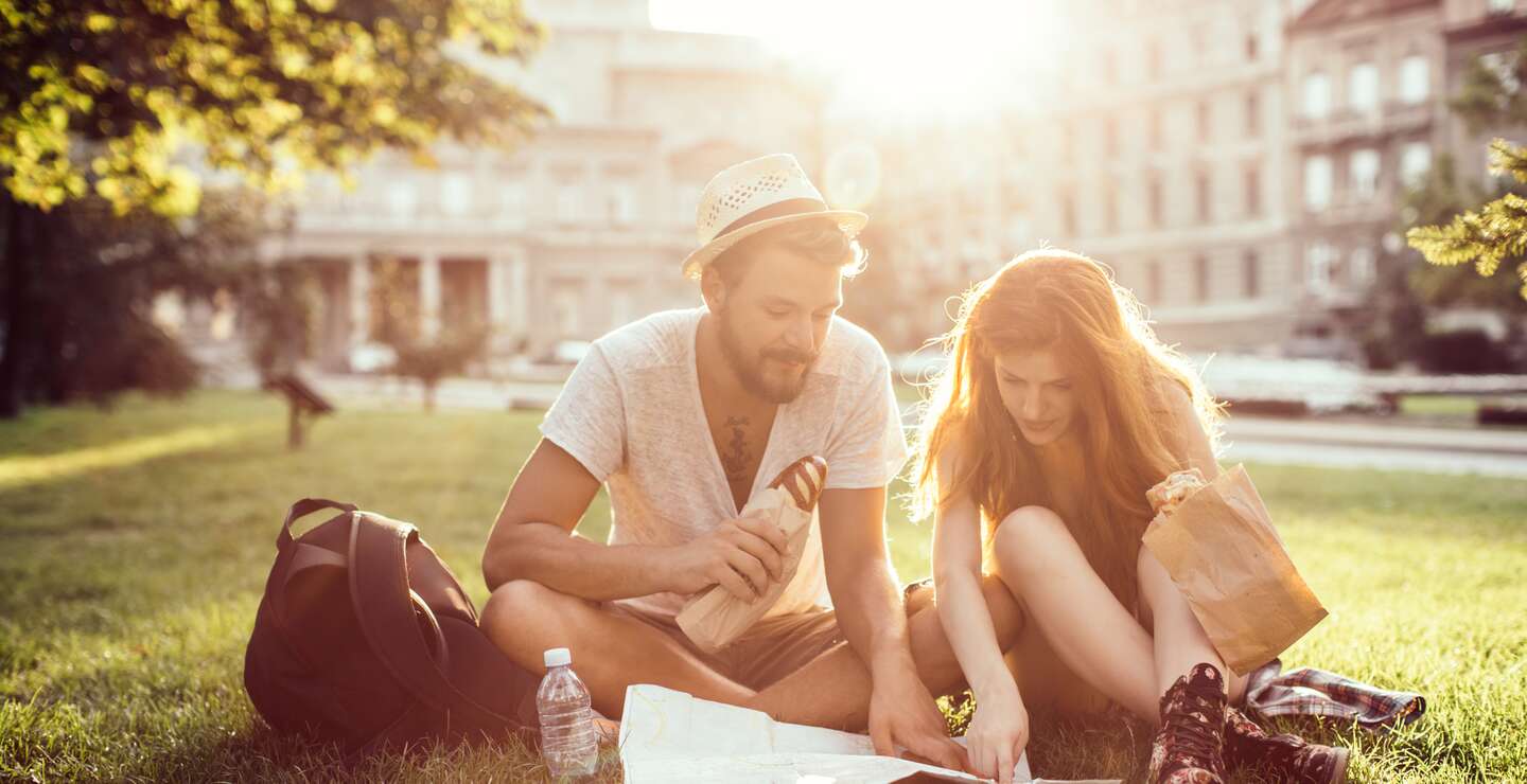 Junges Touristenpaar, das die fremde Stadt erkundet. Auf dem Gras im Park sitzend, frühstückend und Stadtplan lesend. Tragen Freizeitkleidung. | © Gettyimages.com/svetikd