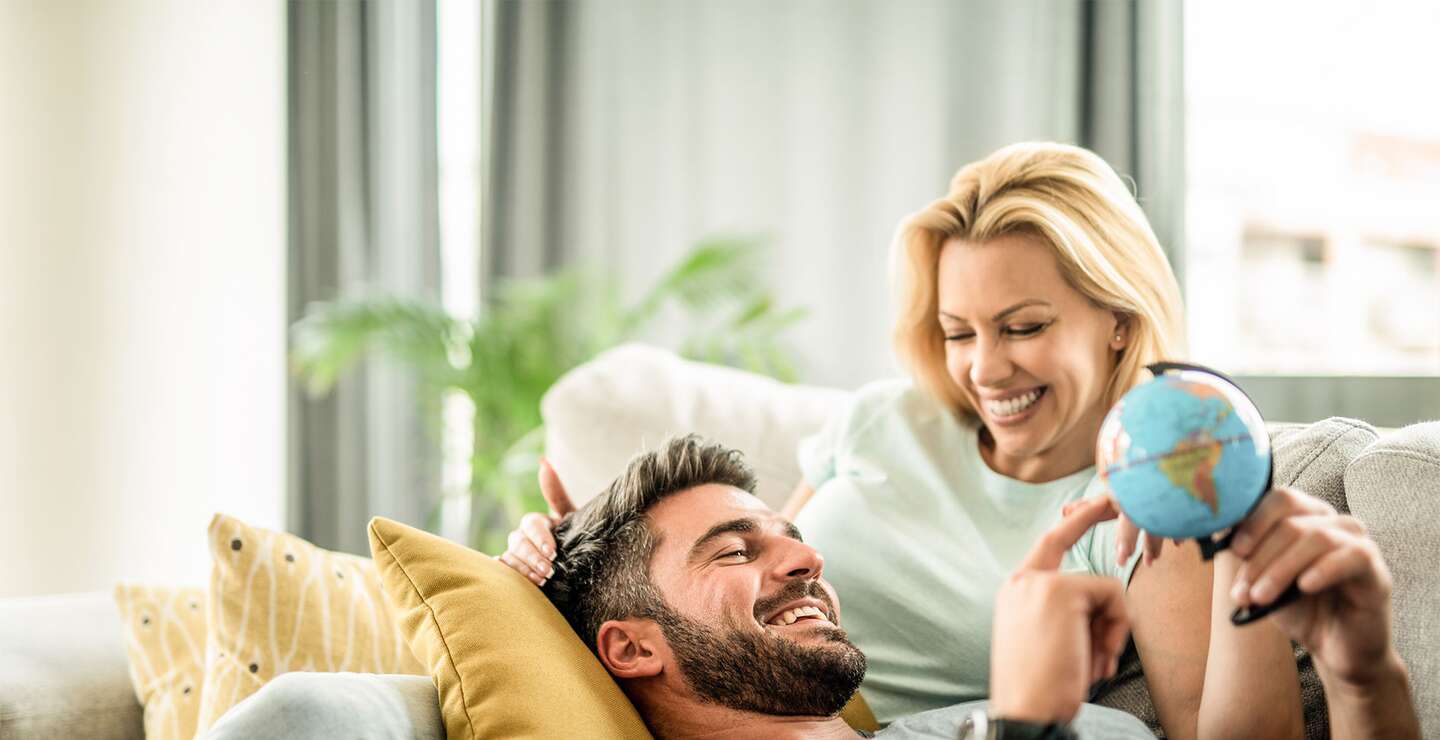 Glückliches junges Paar plant ihre neue Reise. Junger Mann entspannt sich mit seiner Freundin auf dem Sofa und blickt auf den Globus. | © GettyImages.com/DjelicS