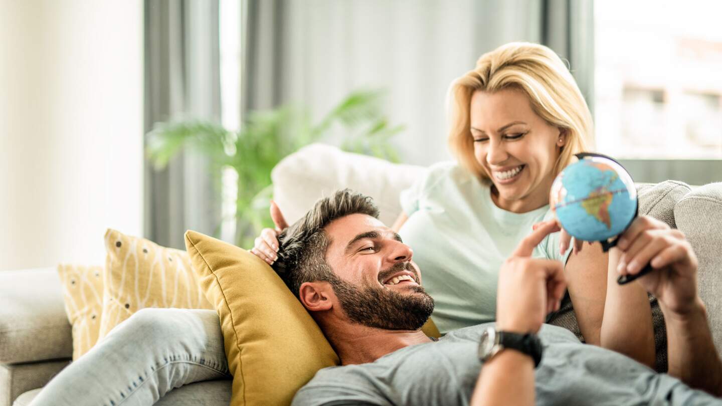 Glückliches junges Paar plant ihre neue Reise. Junger Mann entspannt sich mit seiner Freundin auf dem Sofa und blickt auf den Globus. | © GettyImages.com/DjelicS