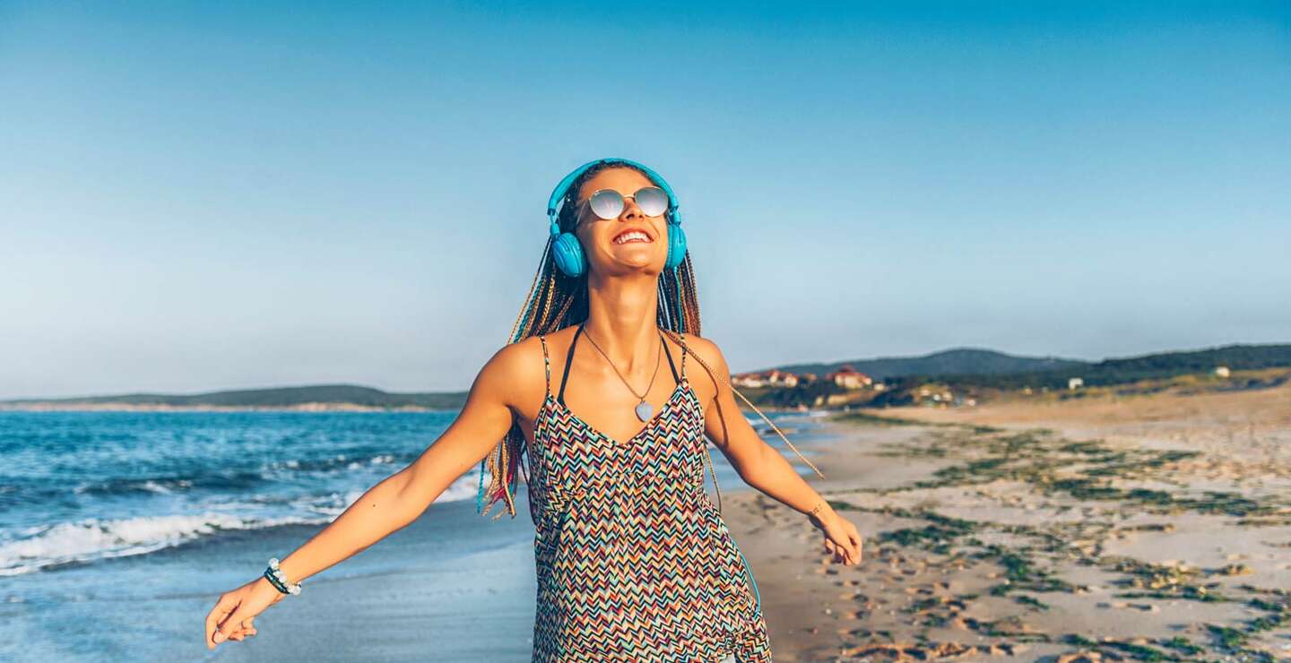 Fröhliche junge Frau mit blauen Kopfhörern, die am Strand Musik hört | © Gettyimages.com/Vladimir Vladimirov