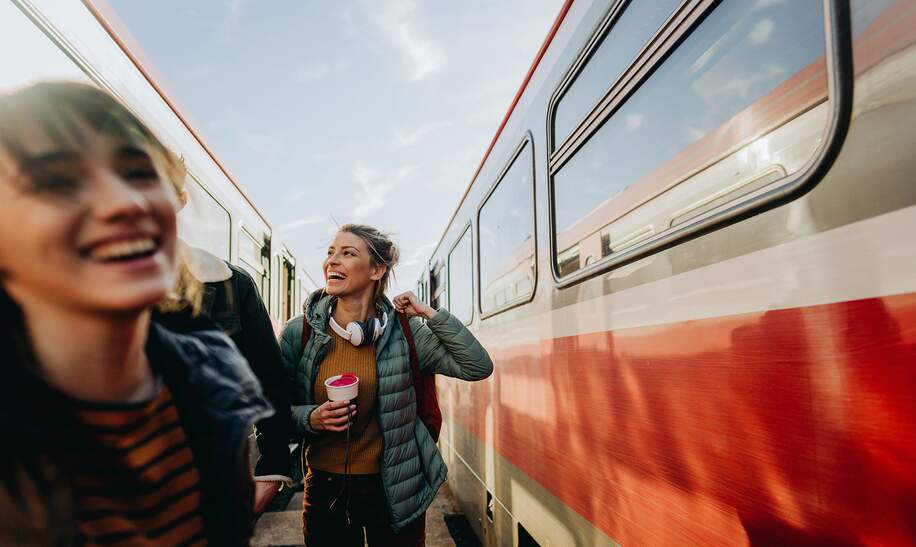 Zwei gut gelaunte, junge Frauen auf dem Bahnsteig zwischen zwei Zügen | © Gettyimages.com/AleksandarNakic