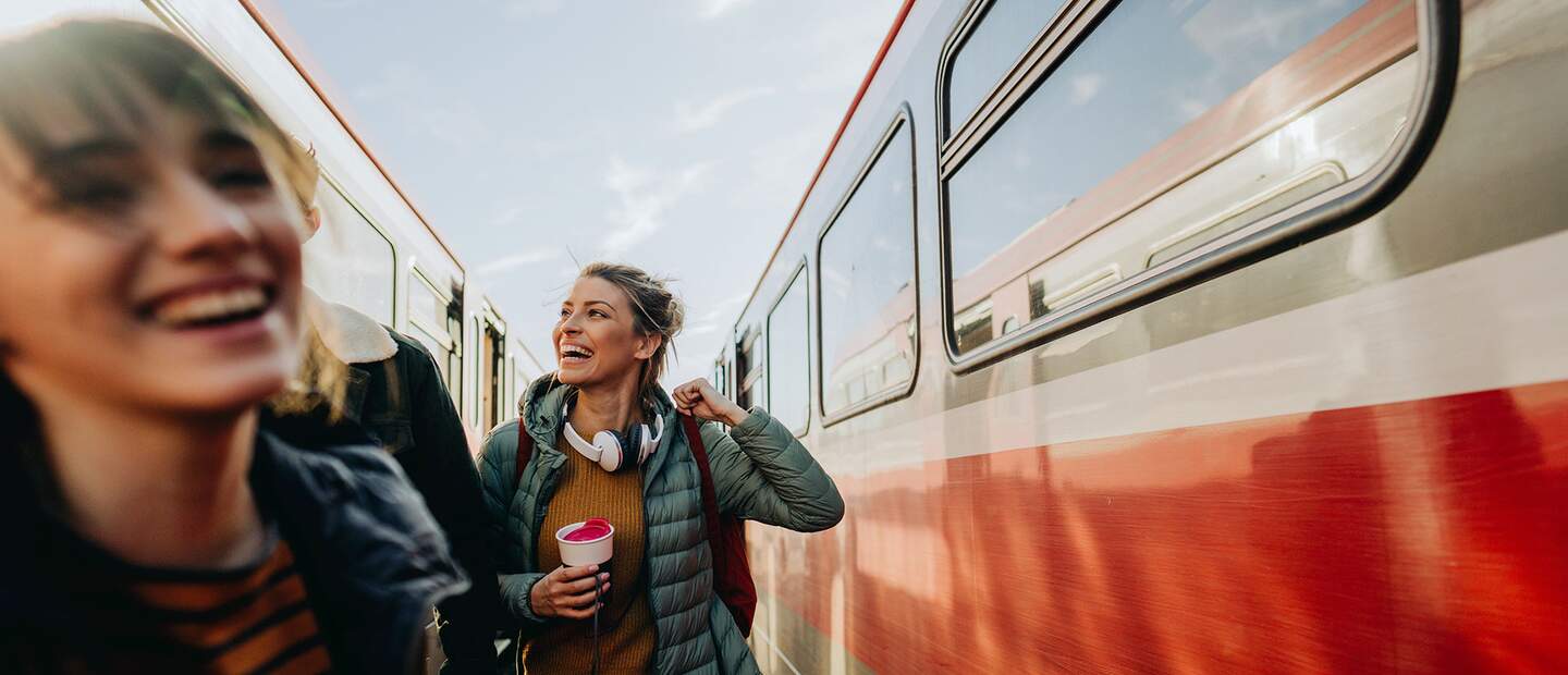 Zwei gut gelaunte, junge Frauen auf dem Bahnsteig zwischen zwei Zügen | © Gettyimages.com/AleksandarNakic