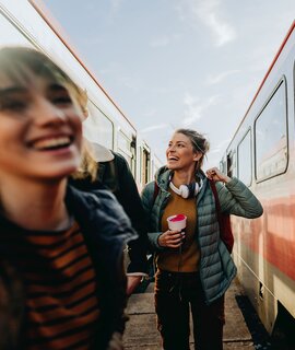 Zwei gut gelaunte, junge Frauen auf dem Bahnsteig zwischen zwei Zügen | © Gettyimages.com/AleksandarNakic