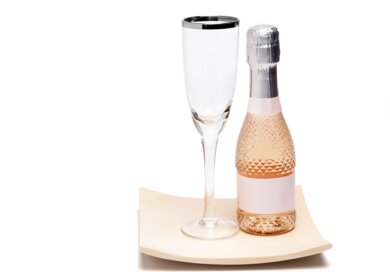 Nahaufnahme einer kleinen Flasche Champagner neben einem edlen Glas mit silbernem Rand auf einer leicht geschwungenen Kiefernholzplatte auf weißem Hintergrund | © Gettyimages.com/PictureSyndicate