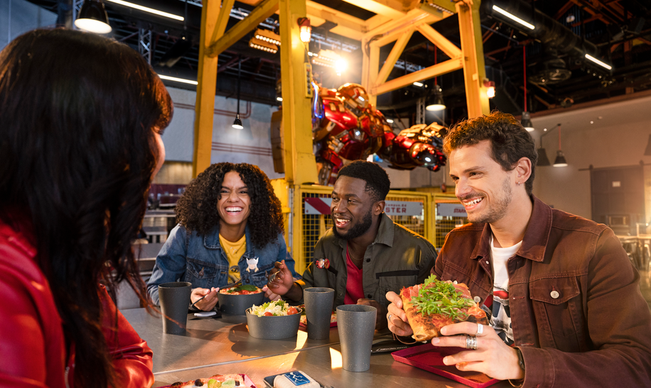 Walt Disney Studios® Park Schnellrestaurant Stark Factory Freunde essen Pizza und Pasta. Im Hintergrund ist Hulkbuster. | © 2024 MARVEL.