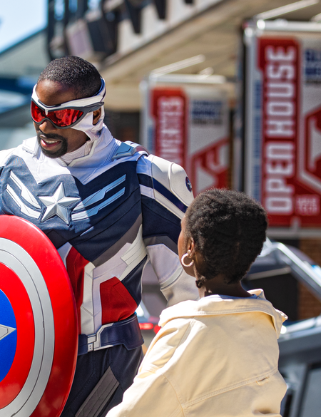 Walt Disney Studios® Park Marvel Avengers Campus Captain America (Sam) mit seinem Schild und zwei Kindern, im Hintergrund der Quinjet | © 2024 MARVEL.