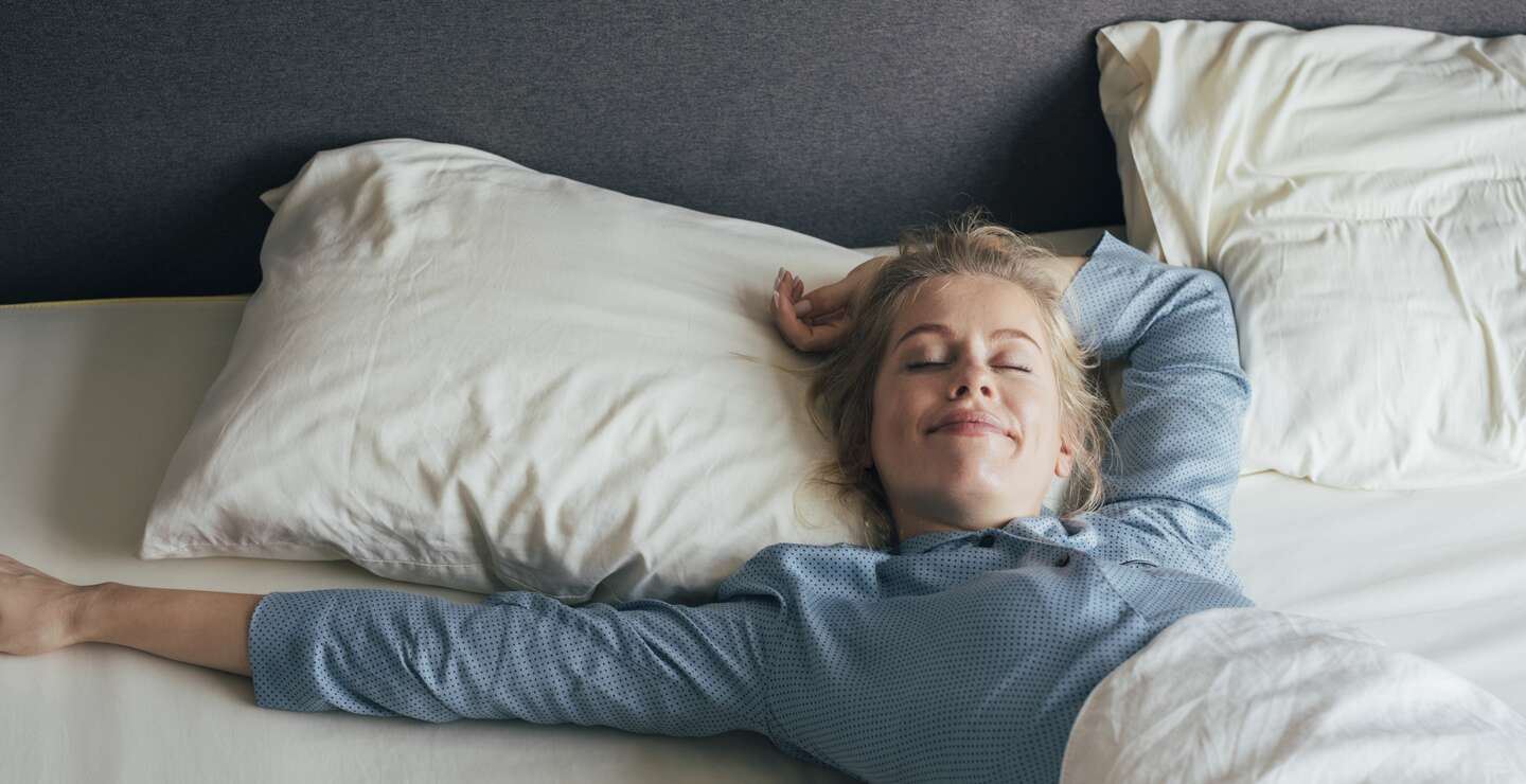 Lächelnde junge Frau, die an einem sonnigen Morgen ihre Arme im Bett streckt | © Gettyimages.com/FreshSplash