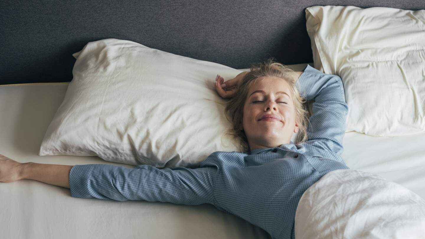 Lächelnde junge Frau, die an einem sonnigen Morgen ihre Arme im Bett streckt | © Gettyimages.com/FreshSplash