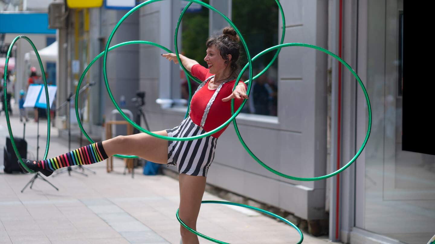 An einem heißen Tag in der Stadt dreht eine Straßenkünstlerin Reifen um ihre Arme und Beine, während sie auf einem Bein balanciert. Sie trägt ein gestreiftes, farbenfrohes Kostüm | © Gettyimages.com/Vladimir Vladimirov