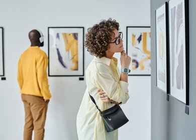Junge Frau mit Brille untersucht moderne Kunst in der Galerie mit anderen Besuchern | © Gettyimages.com/AnnaStills
