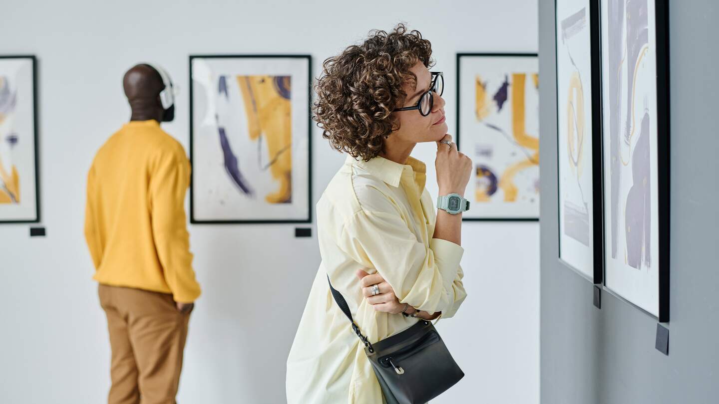 Junge Frau mit Brille untersucht moderne Kunst in der Galerie mit anderen Besuchern | © Gettyimages.com/AnnaStills