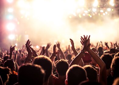 Publikum mit erhobenen Händen bei einem Musikfestival und Lichtern, die von oben auf die Bühne strömen | © Gettyimages.com/bernardbodo