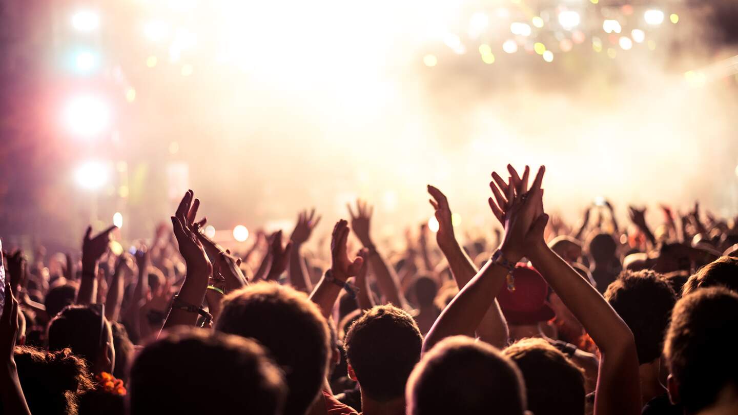 Publikum mit erhobenen Händen bei einem Musikfestival und Lichtern, die von oben auf die Bühne strömen | © Gettyimages.com/bernardbodo