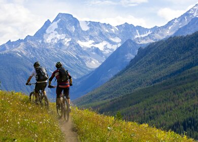 Zwei Radfahrer fahren über einen Berg in British Columbia, Kanada und sind dabei von hinten zu sehen, vor ihnen liegt ein verschneiter Berg | © GettyImages.com/GibsonPictures