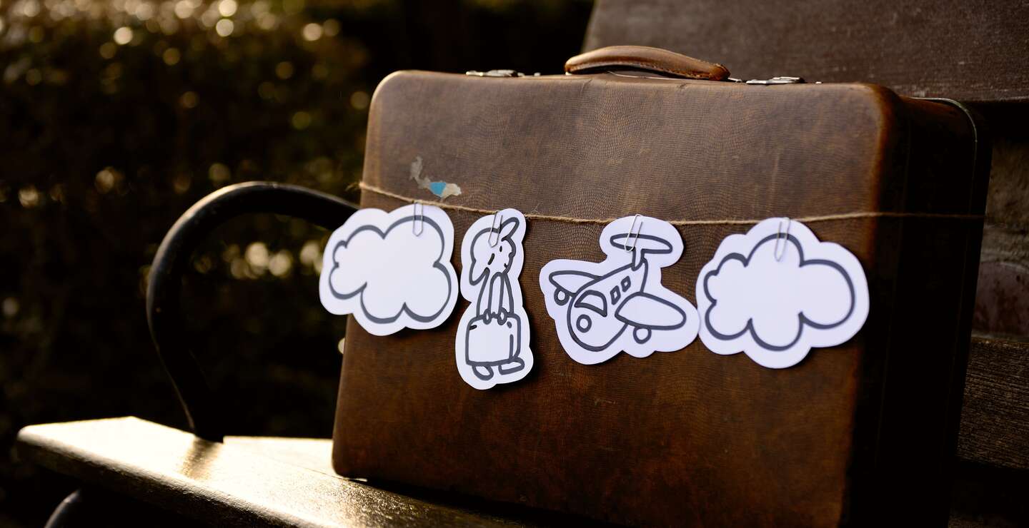 Braune Aktentasche steht auf Bank und hat ausgeschnittenes, bemaltes papier mit Reisendem, Flugzeug und Wolken an einer Schnur befestigt umgebunden | © Pixabay/congerdesign