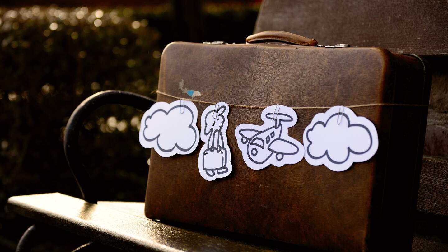 Braune Aktentasche steht auf Bank und hat ausgeschnittenes, bemaltes papier mit Reisendem, Flugzeug und Wolken an einer Schnur befestigt umgebunden | © Pixabay/congerdesign