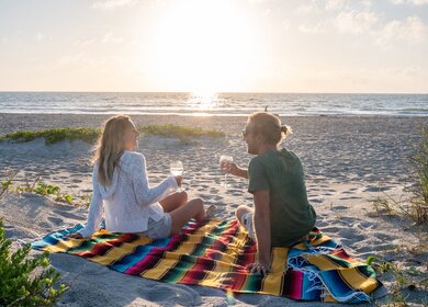 Zwei Personen teilen sich den Moment bei Sonnenuntergang, während sie sich auf einer Decke mit einem Glas Wein im Freien am Strand entspannen | © Gettyimages.com/swissmediavision