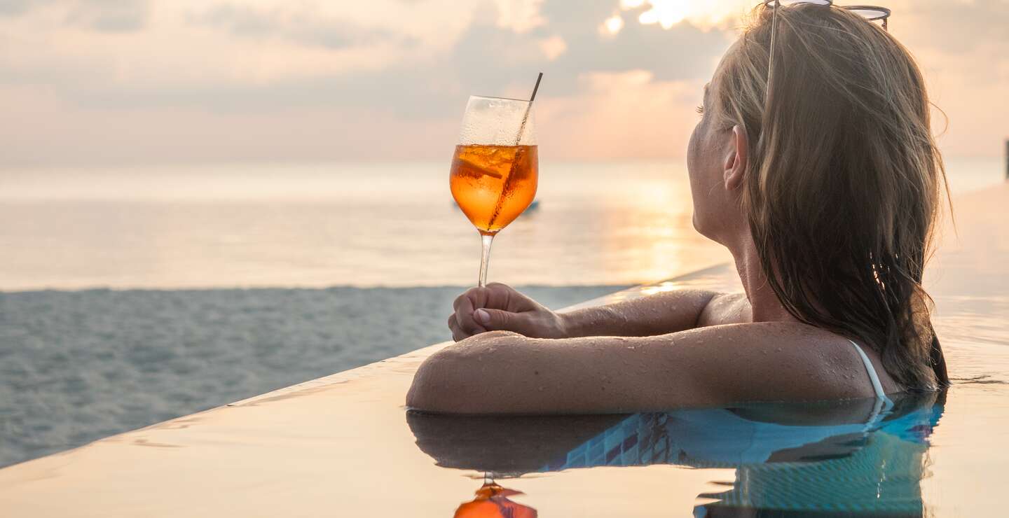 Frau trinkt Cocktail bei Sonnenuntergang in einem Infinity-Pool | © Gettyimages.com/Mystockimages