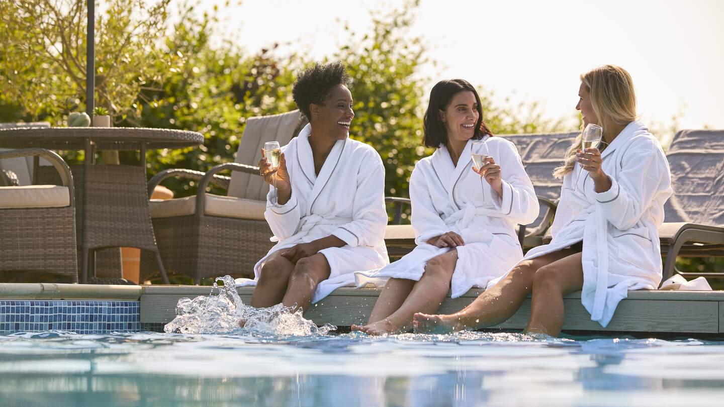 Gruppe von Freundinnen, die Bademäntel tragen, sitzen mit den Füßen im Pool und trinken Champagner am Spa-Tag | © Gettyimages.com/monkeybusinessimages