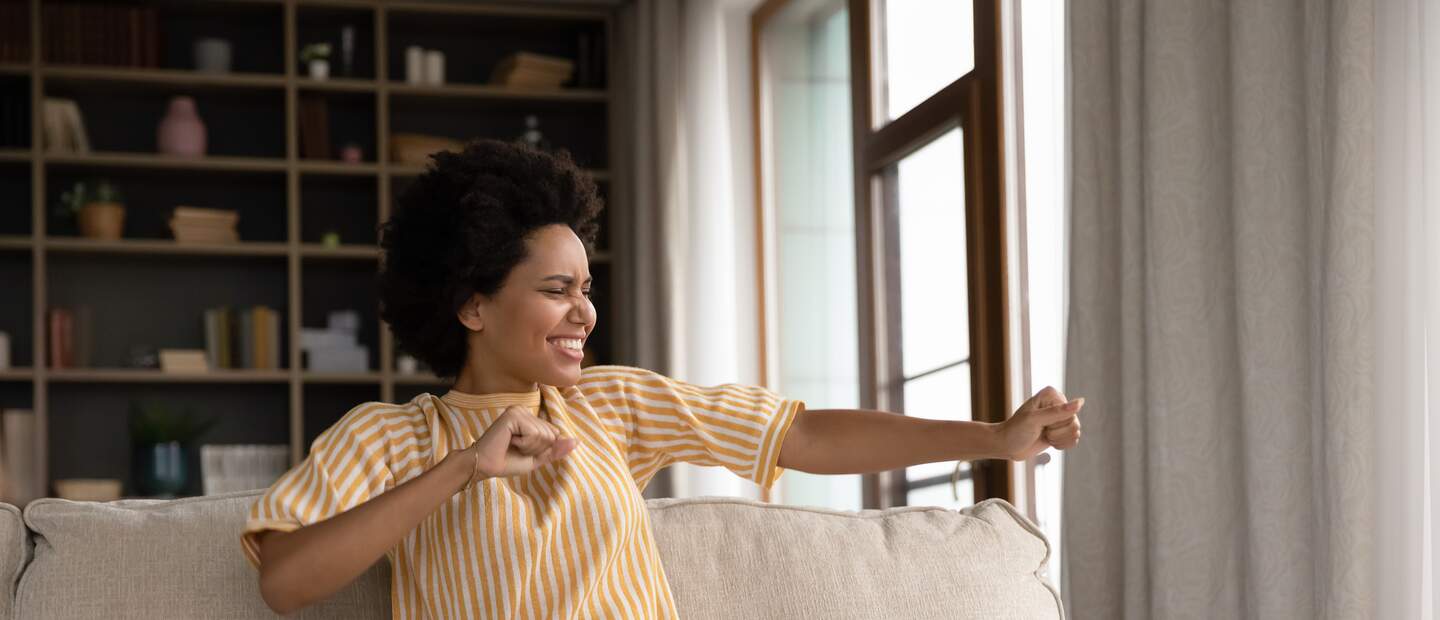 Junge Frau arbeitet von zu Hause | © Gettyimages.com/fizkes  junge afroamerikanische Frau, die zu Hause am Computer arbeitet, den letzten Arbeitstag feiert, Spass allein im Wohnzimmer hat | © Gettyimages.com/fizkes